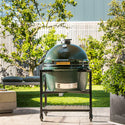 Large green ceramic grill on a stand outdoors with trees and a building in the background