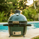 Big Green Egg grill by a pool with blurred greenery in the background