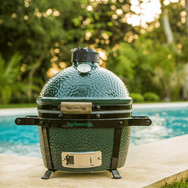 Big Green Egg grill by a pool with blurred greenery in the background