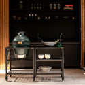 Outdoor kitchen setup with a large green grill on a cart, shelves with cookware in the background.