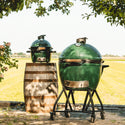 Two green Kamado-style grills outdoors with a wooden barrel and field background.