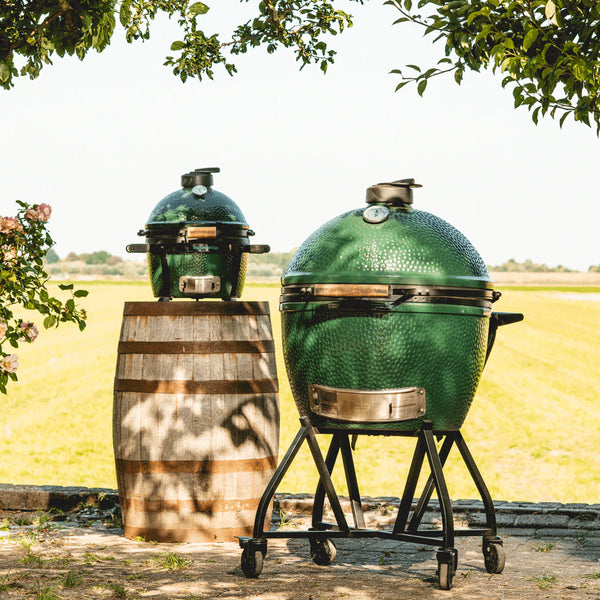 Two green Kamado-style grills outdoors with a wooden barrel and field background.